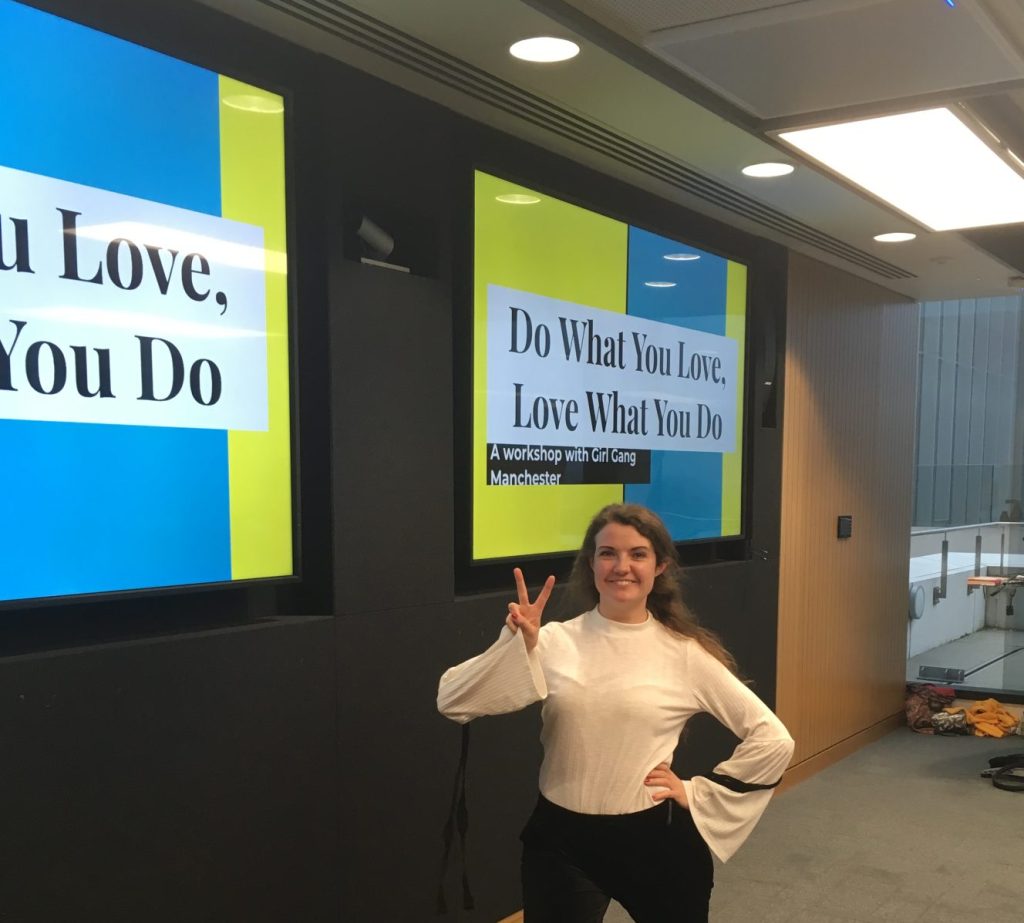 A person stands smiling and making a peace sign in front of two large screens displaying the text "Do What You Love, Love What You Do. A workshop with Girl Gang Manchester." The environment appears to be a modern indoor space.