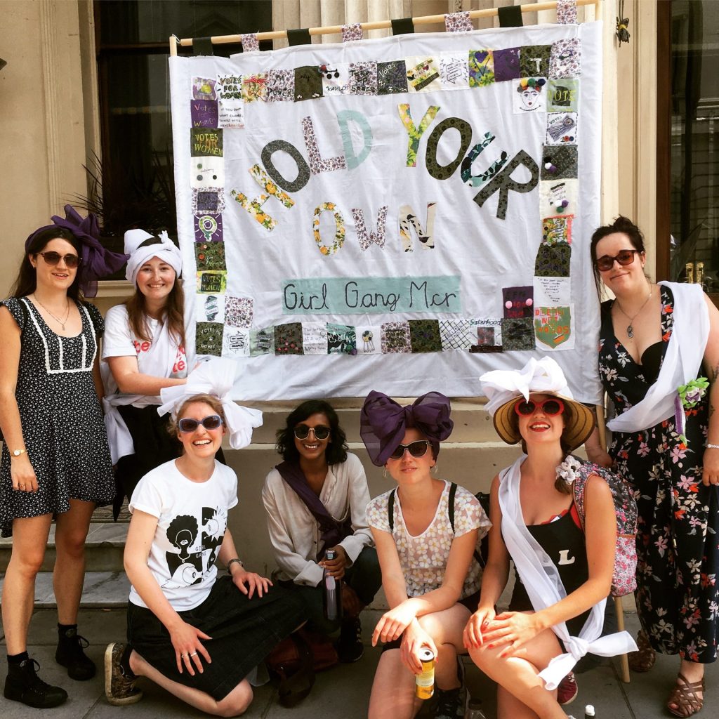 A group of women pose in front of a banner that reads "HOLD YOUR OWN Girl Gang Mer." They wear white sashes and purple bows, with smiles and sunglasses. Some are seated while others stand on steps outside a building.