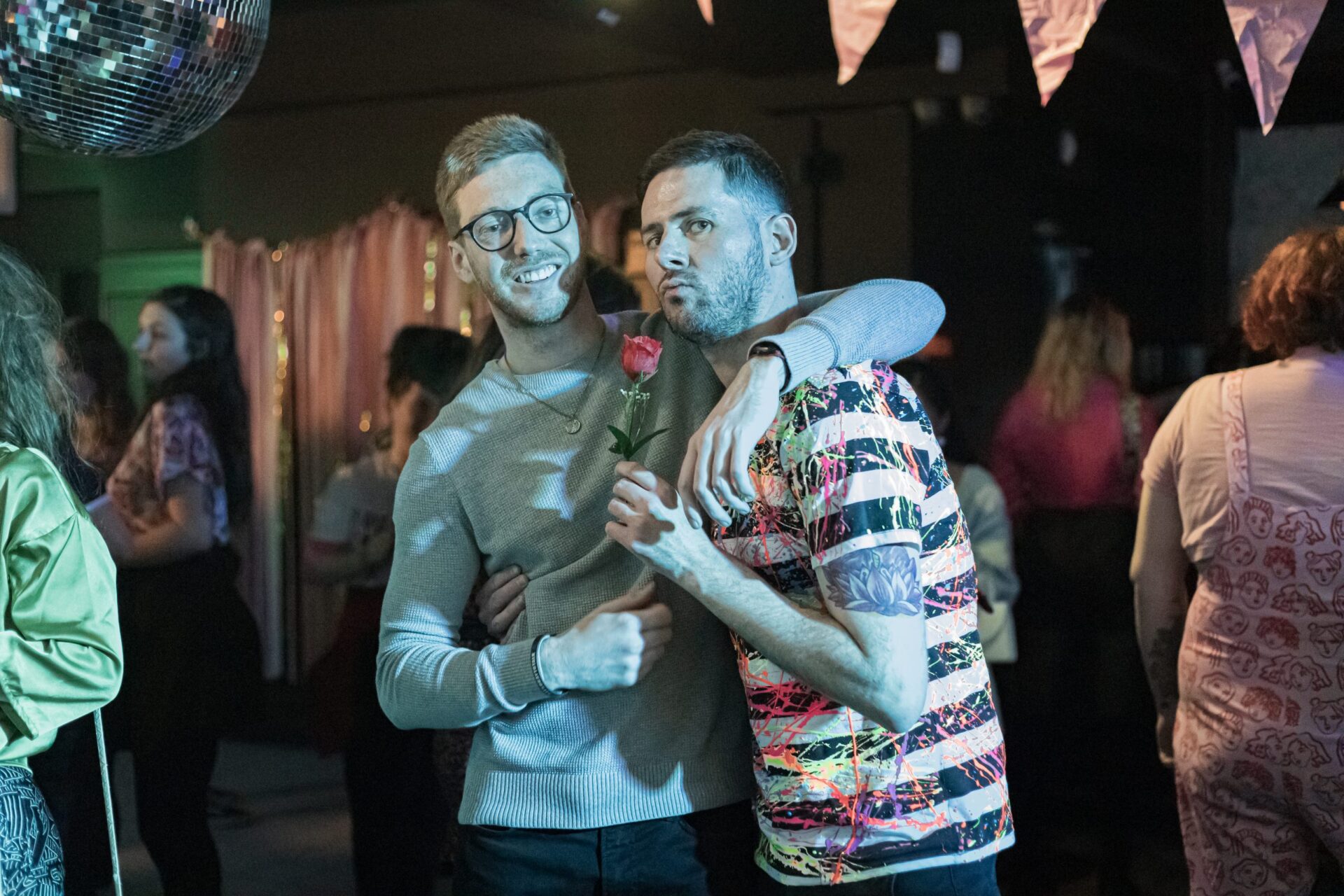 2 men embracing each other at a party, one holding a rose