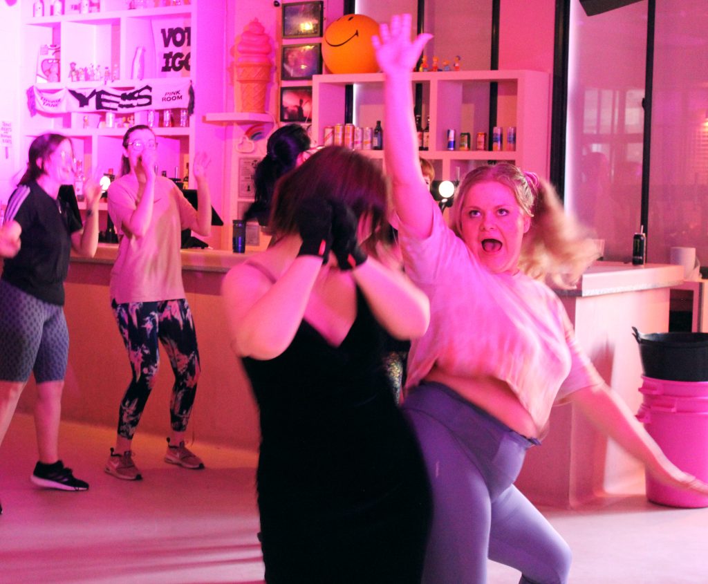 A group of people dance energetically in a pink-lit room. One person in the foreground raises their arm enthusiastically while another dances beside them. Shelves with assorted drinks and decor are in the background.