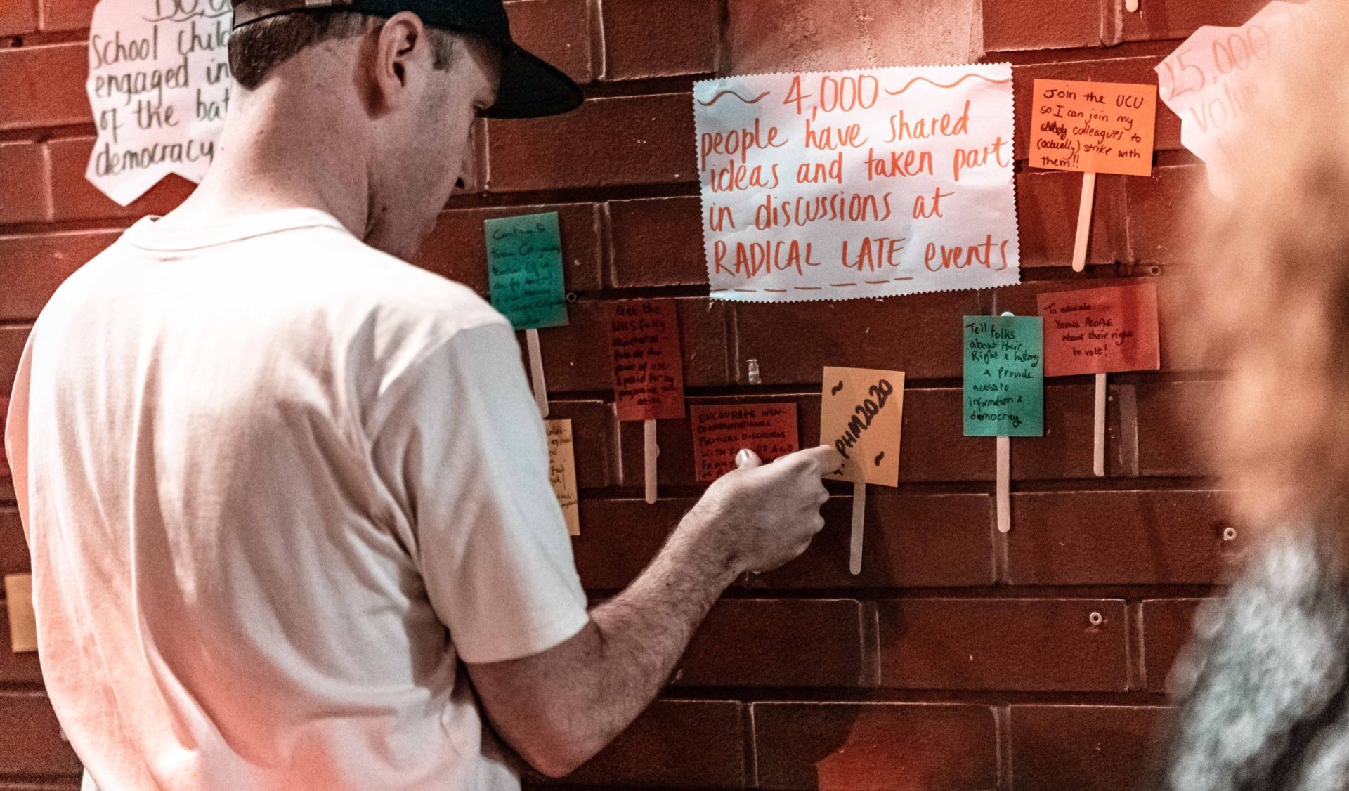 Person in white t-shirt and baseball hat pinning a post it note to a brick wall with many other post-it notes attached to