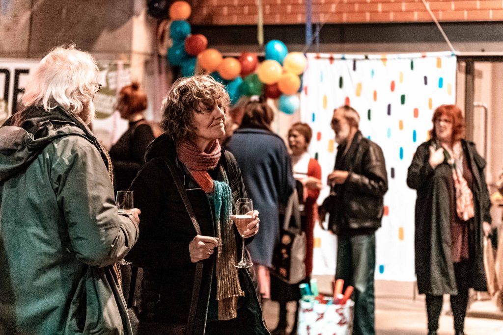 2 older aged people stood holding champagne glasses at a gathering with balloons and decorations in the background