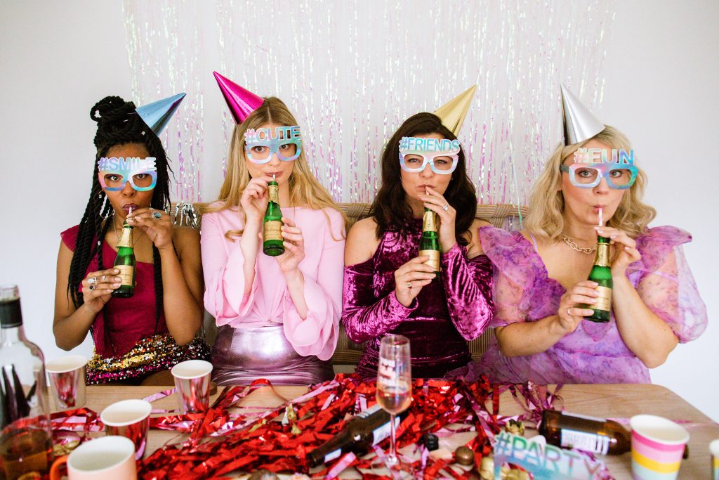 4 people sat in a row dressed up in pink and red clothes, sipping champagne, wearing novelty hashtag glasses and party hats, surrounded by party items