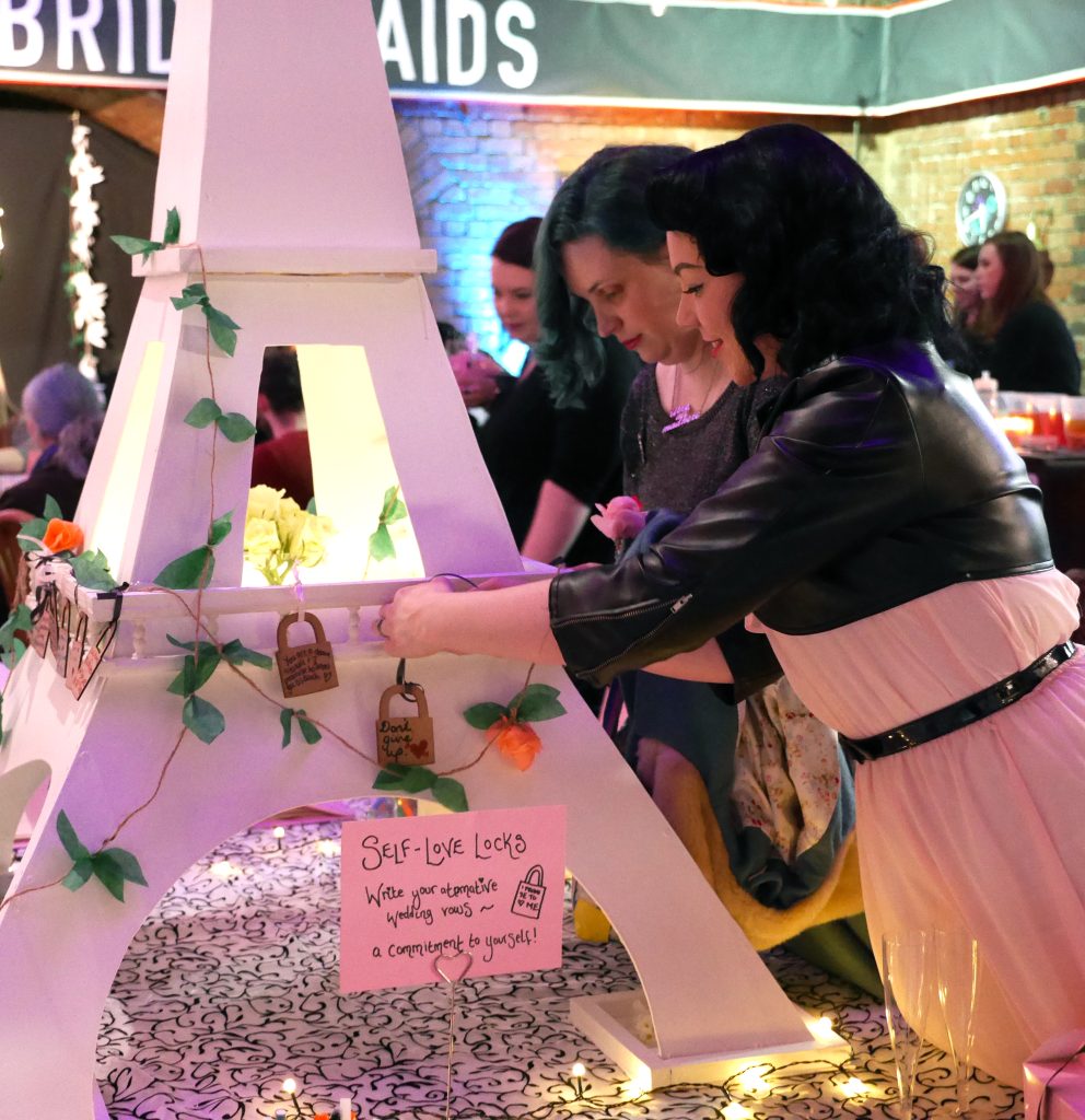 Two people attach locks to a white Eiffel Tower model at an event. A sign reads "Self-Love Locks" encouraging attendees to write positive messages. The backdrop shows festive lighting and a banner.