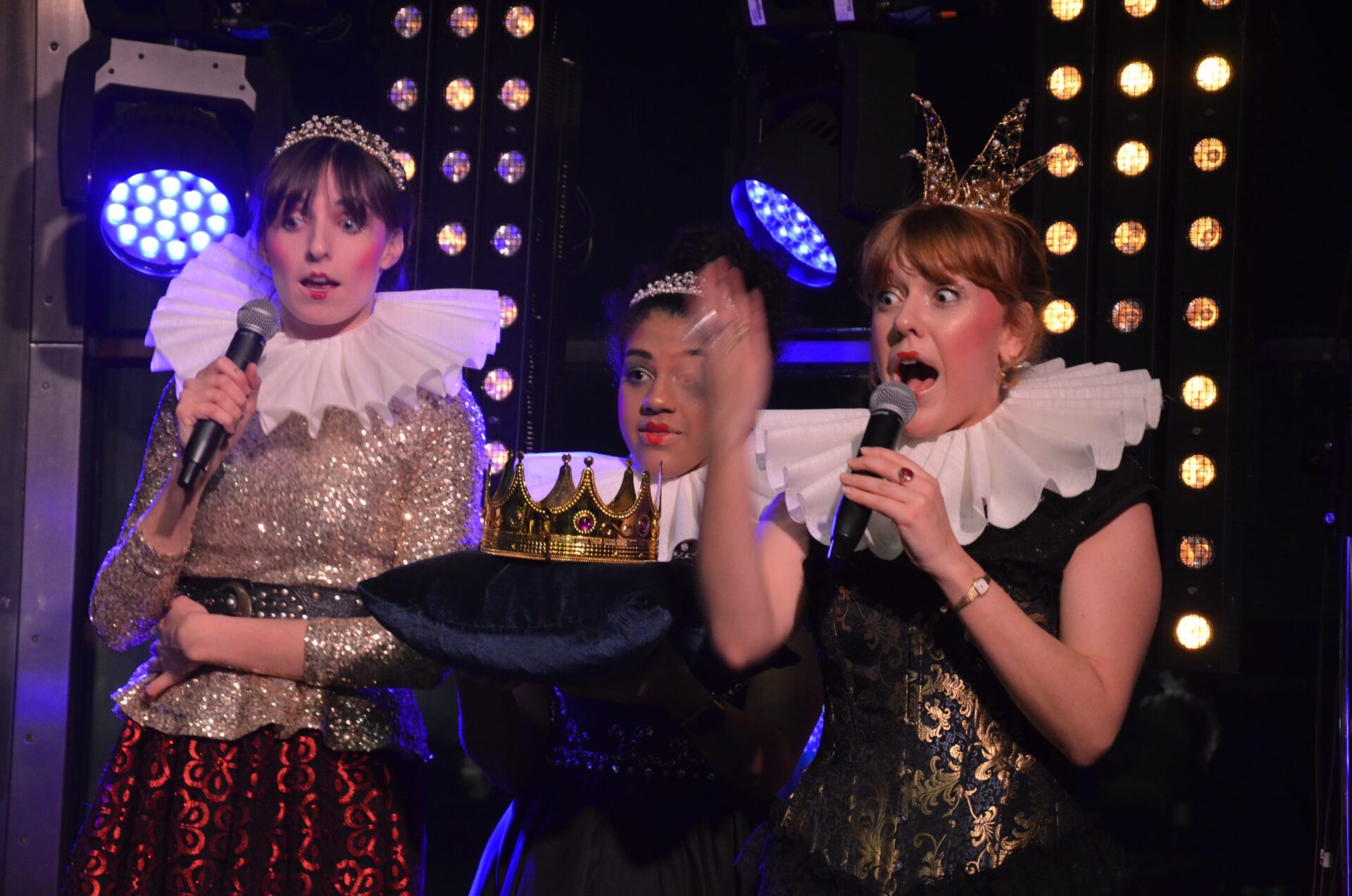 3 people dressed in period royalty costumes, one holding a crown on a cushion and 2 talking expressively into mics, stage lights behind them