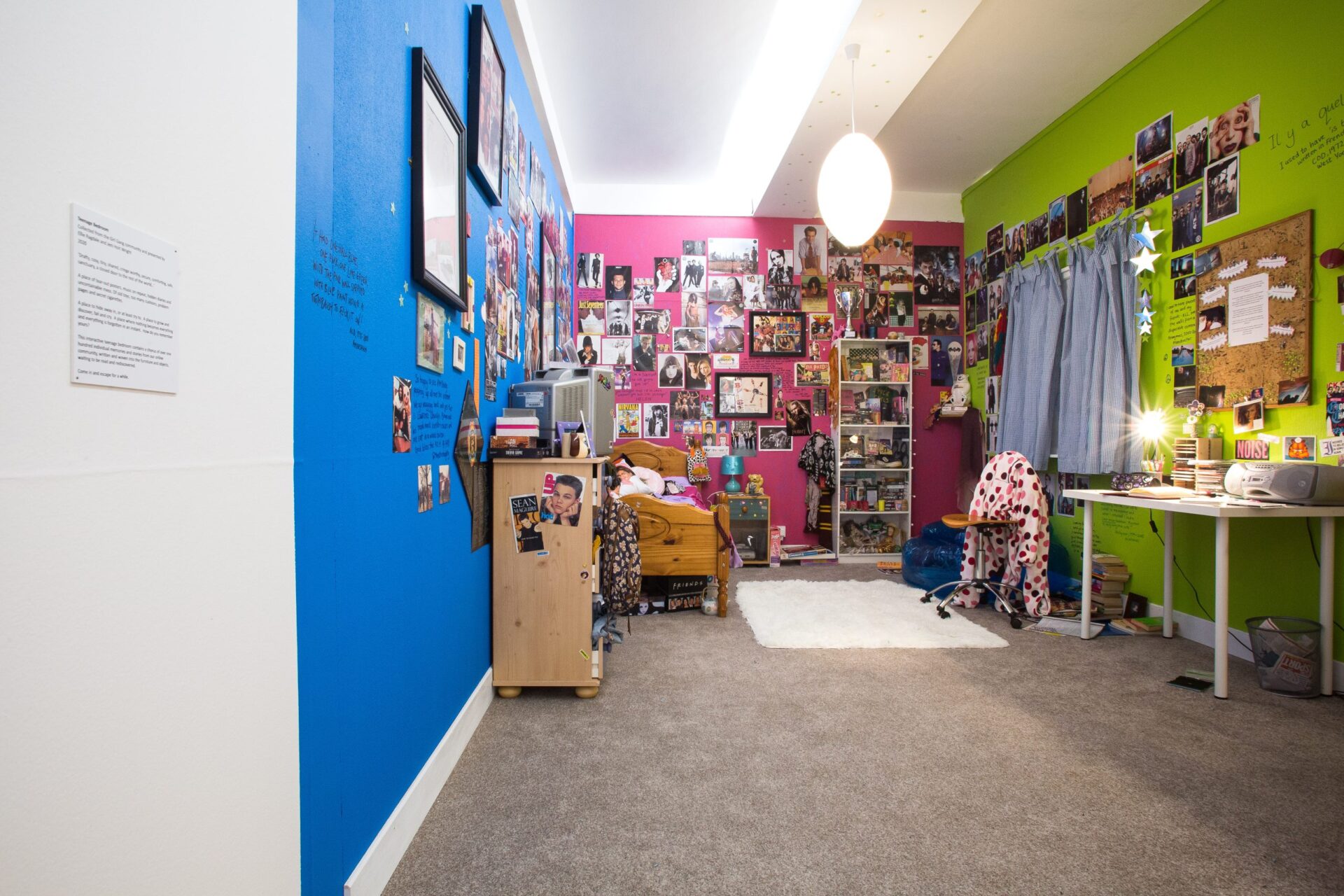 An exhibition installation piece of a stereotypical teenage bedroom with blue pink and green walls with loads of posters and images stuck to the walls