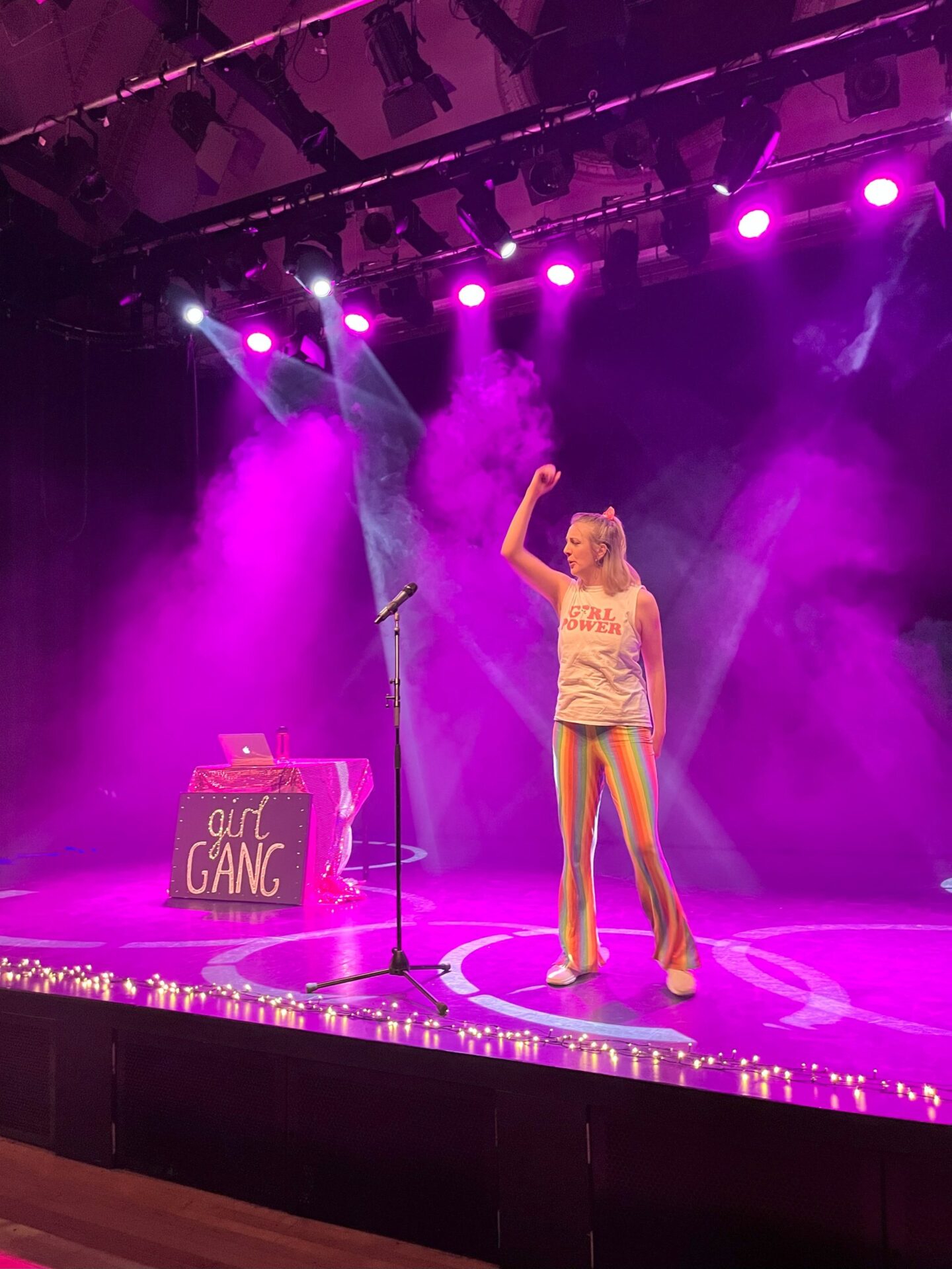 Woman wearing multi-coloured stripey trousers and Girl Power T-shirt on a purple lit stage in a power stance with arm up