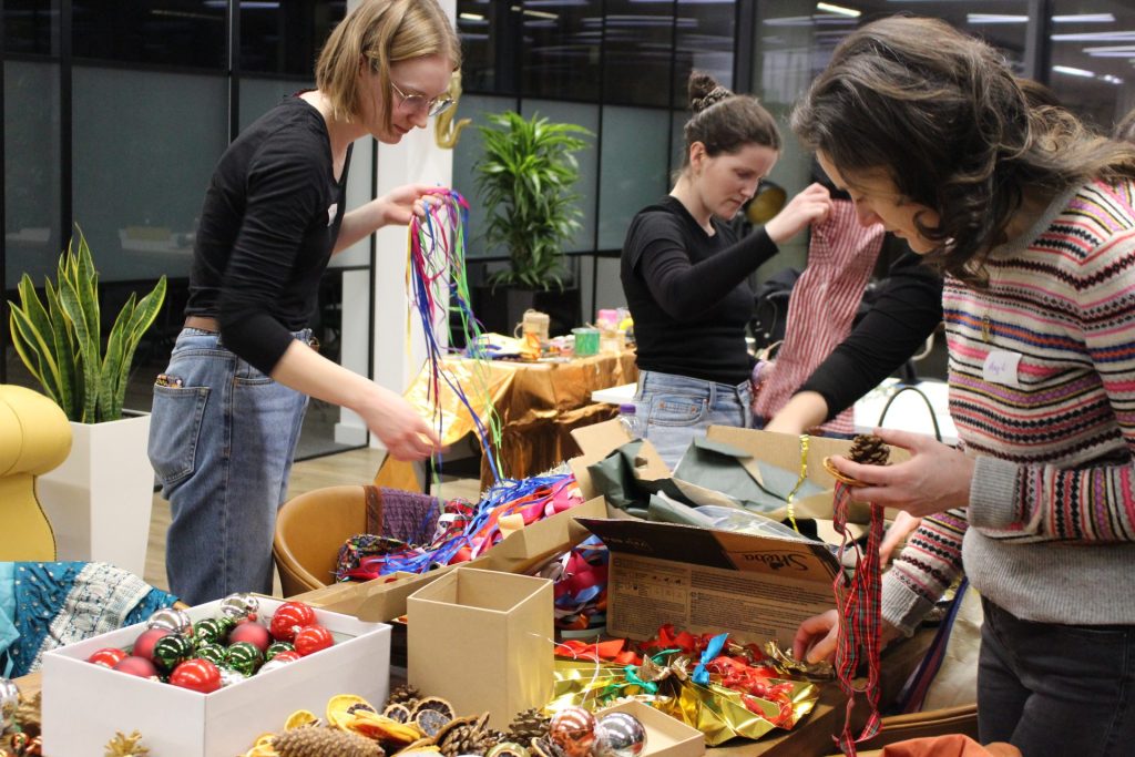 People at a table crafting with colorful ribbons, ornaments, and pinecones. They are in a bright room with large windows and plants, creating festive decorations.