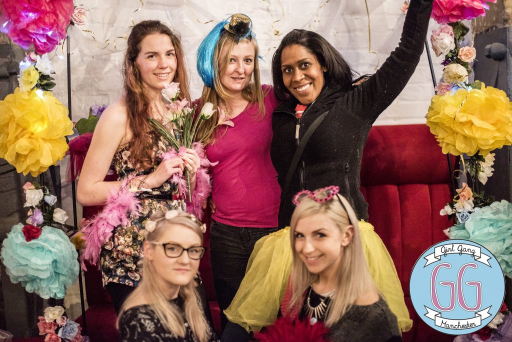 Five women are gathered in front of a festive backdrop with colorful tissue paper flowers. They wear playful accessories like boas and tiaras. A logo reading "Girl Gang Manchester" is in the corner. They are smiling and posing for the camera.