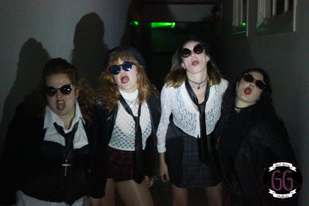 Four people wearing sunglasses, dressed in edgy, gothic-inspired outfits with ties and lace shirts, pose playfully in a dimly lit hallway. A neon green light is visible in the background, and a circular logo is on the bottom right corner.