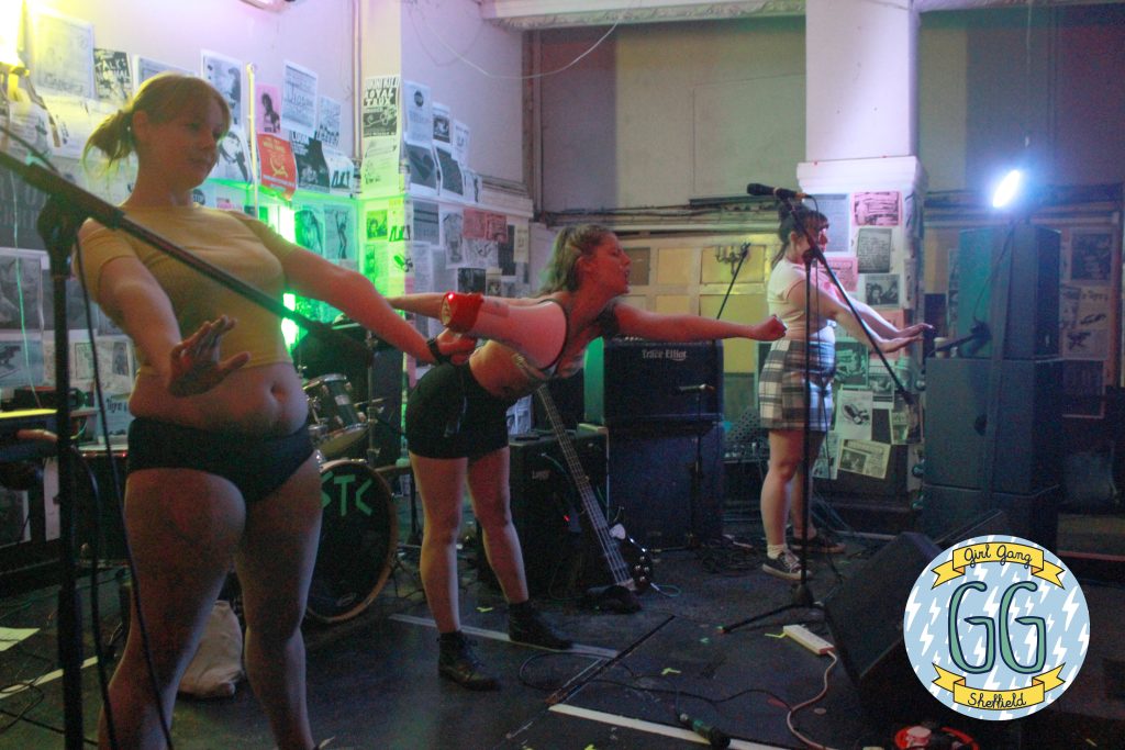 Three performers on stage, with one holding a microphone and two others striking expressive poses. The backdrop is filled with posters, and instruments are visible. The scene is illuminated by colorful lights.