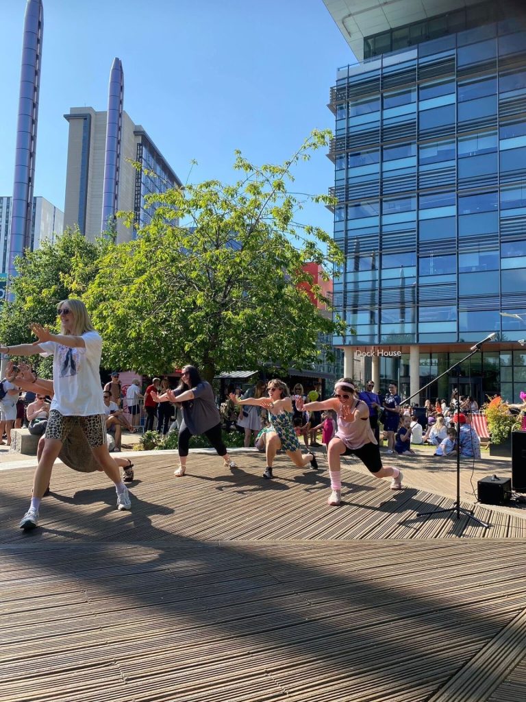 A group of people is performing a dance routine on an outdoor stage surrounded by a crowd. The stage is situated near a modern glass building and several tall structures. Green trees and a clear blue sky complete the lively, urban setting.