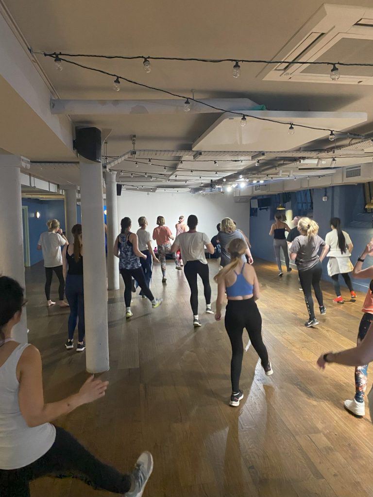 Image of group of women from behind, they are moving their bodies with one leg in the air, in a large indoor space