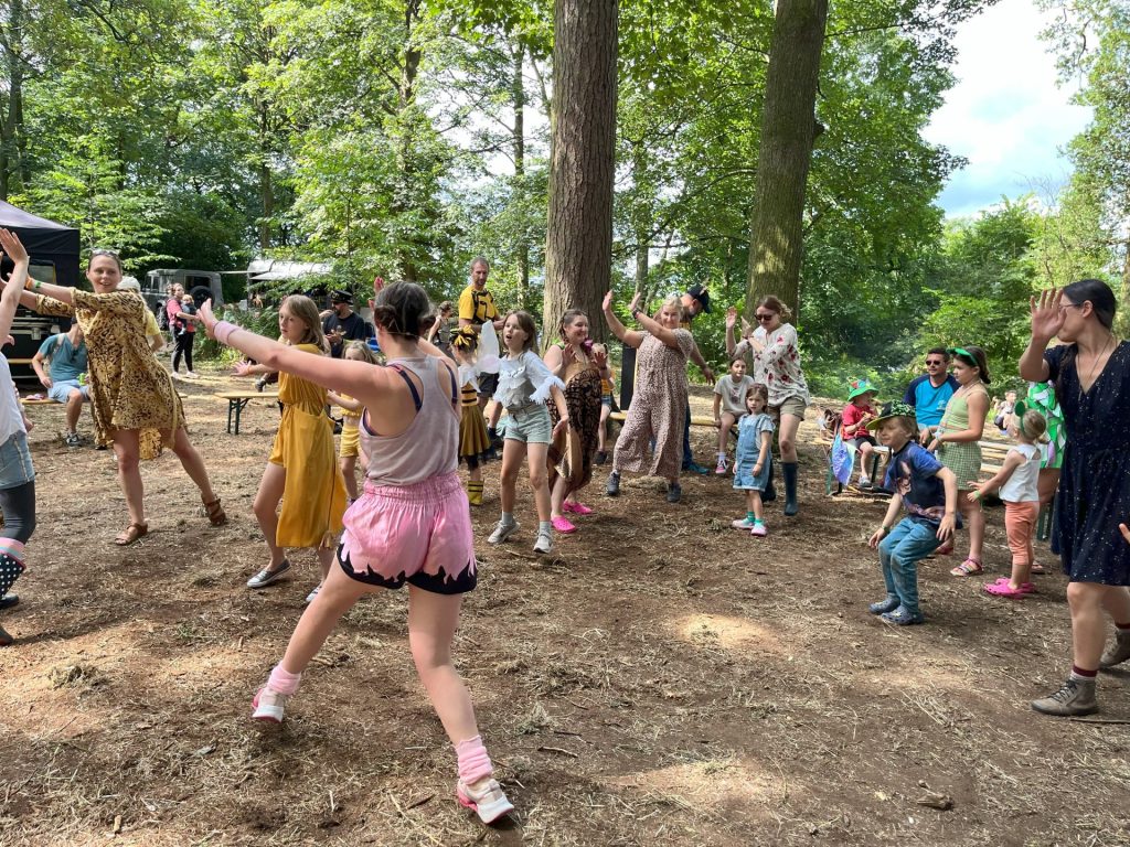 A lively outdoor scene in a forested area features people of various ages dancing energetically. Children and adults in colorful clothing are moving joyfully under the trees, with picnic tables and a tent visible in the background.