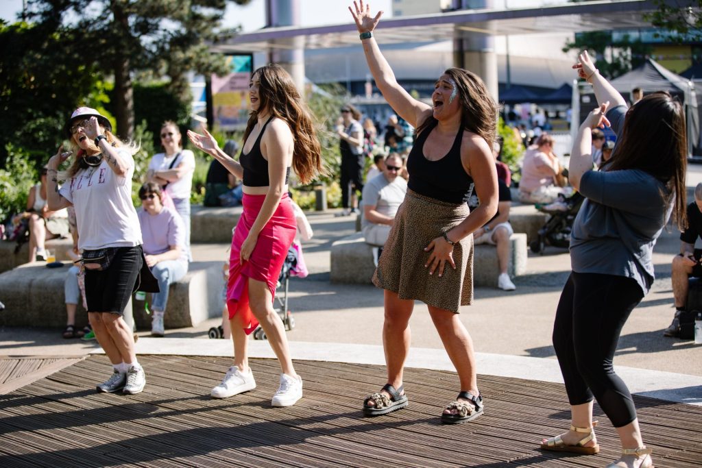 A group of four people enthusiastically dancing on a wooden deck outdoors. The participants are smiling and appear to be enjoying a sunny day. Other people are seen sitting and walking in the background, contributing to a lively and fun atmosphere.