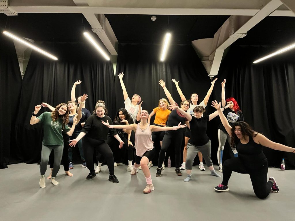 A group of people, some dressed in dance attire, pose energetically in a studio with dark curtains and overhead lighting. They exhibit various dance stances, with arms raised, legs bent, and expressive postures, appearing joyful and enthusiastic.