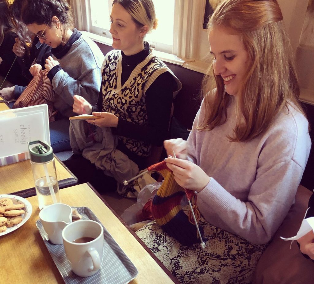 Three women sit at a table, smiling and knitting colorful items. There are two mugs, a small glass pot on a tray, and cookies on a plate. The atmosphere is cozy and casual.