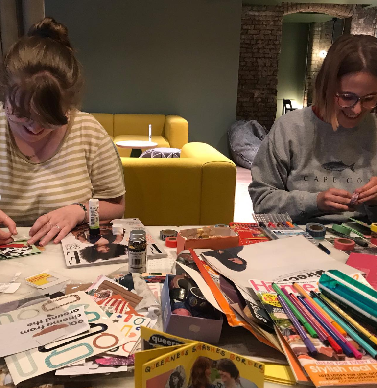 Two people sitting at a table filled with art supplies like markers, magazines, and scissors. They're smiling while crafting, with a yellow couch and a stone wall in the background.