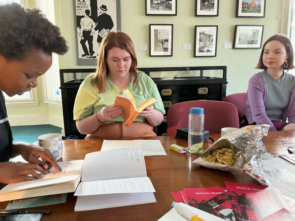 Three people sitting around a wooden table with books, papers, and snacks. One person is writing, another is reading, and a third is listening. Walls have framed pictures. Cups and a water bottle are on the table.