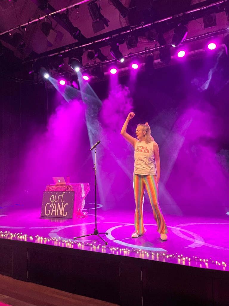 A person performs on stage under pink and purple lighting, wearing a "girl gang" shirt and colorful striped pants. Behind them is a table with a sign that says "girl gang," surrounded by small lights. A microphone is on the stage.