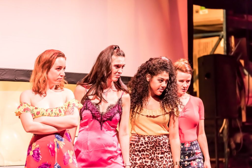 Four women are standing close together on stage, looking towards the right. They are dressed in colorful outfits, with one wearing a floral skirt and others in bright tops or dresses. The background shows part of a screen and stage lights.