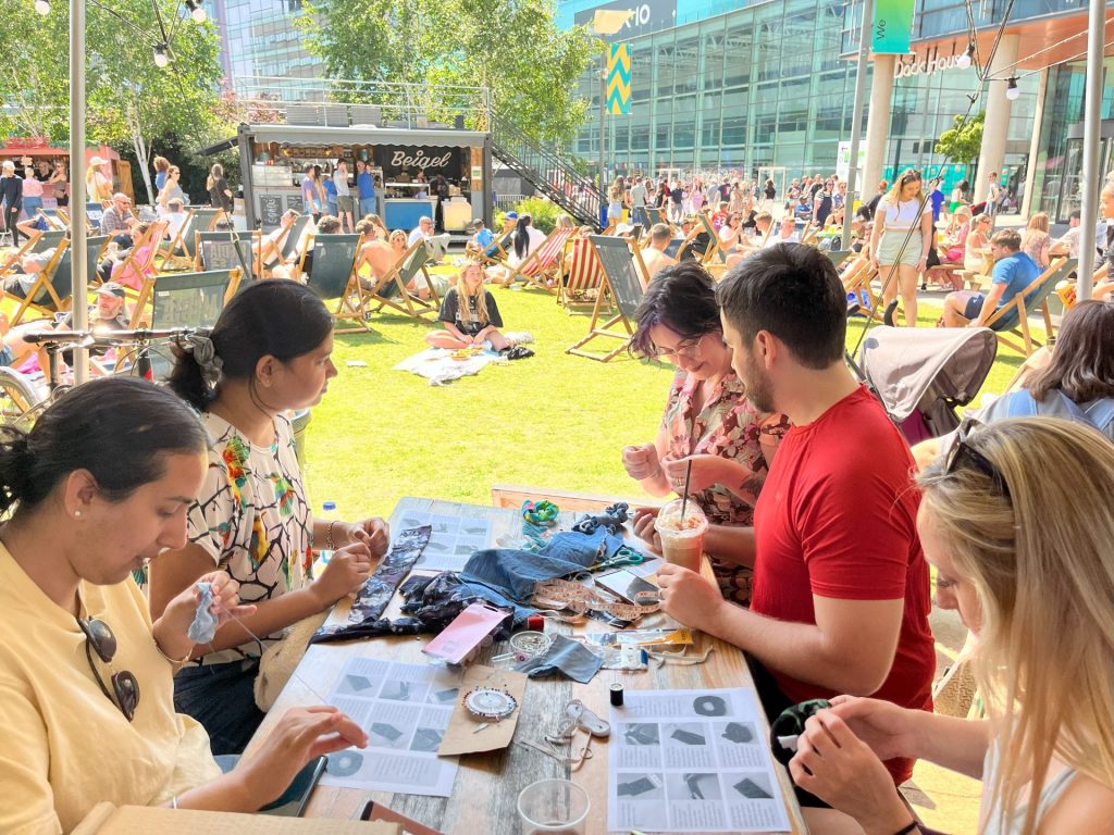 A group of people sit around a table outdoors, engaged in crafting activities. The park area is bustling with people relaxing on deck chairs under sunny skies. A mix of greenery and modern buildings can be seen in the background.