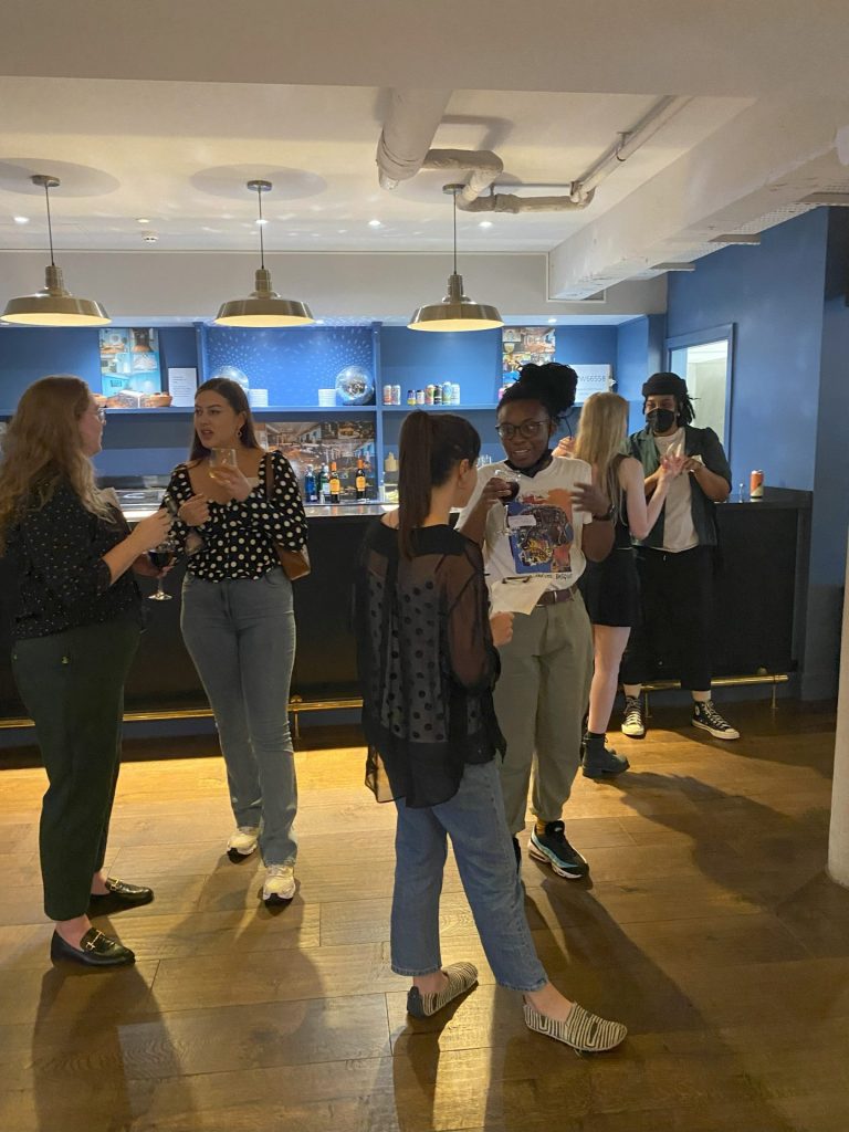 group of people stood in front of a bar talking with one another holding drinks