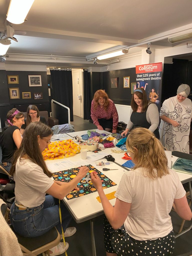 A group of people are gathered around a table in a room, working on crafting projects with fabric and sewing materials. The room has black and white photos on the walls and a poster for Colosseum Theatre.