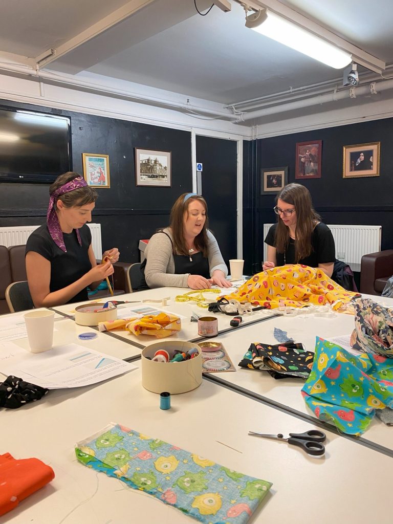 Three women sit around a table engaged in sewing activities. Fabrics, threads, and sewing tools are spread on the table. They appear focused and collaborative in a room decorated with framed pictures on the wall.