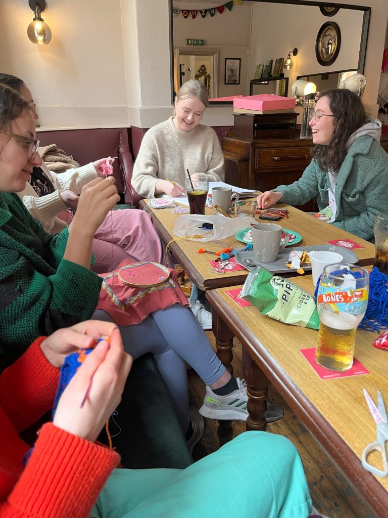 A group of people sitting in a cozy room, engaged in crafting activities around a table. Various supplies like yarn and embroidery hoops are scattered across the table. They appear to be enjoying themselves, smiling and chatting.