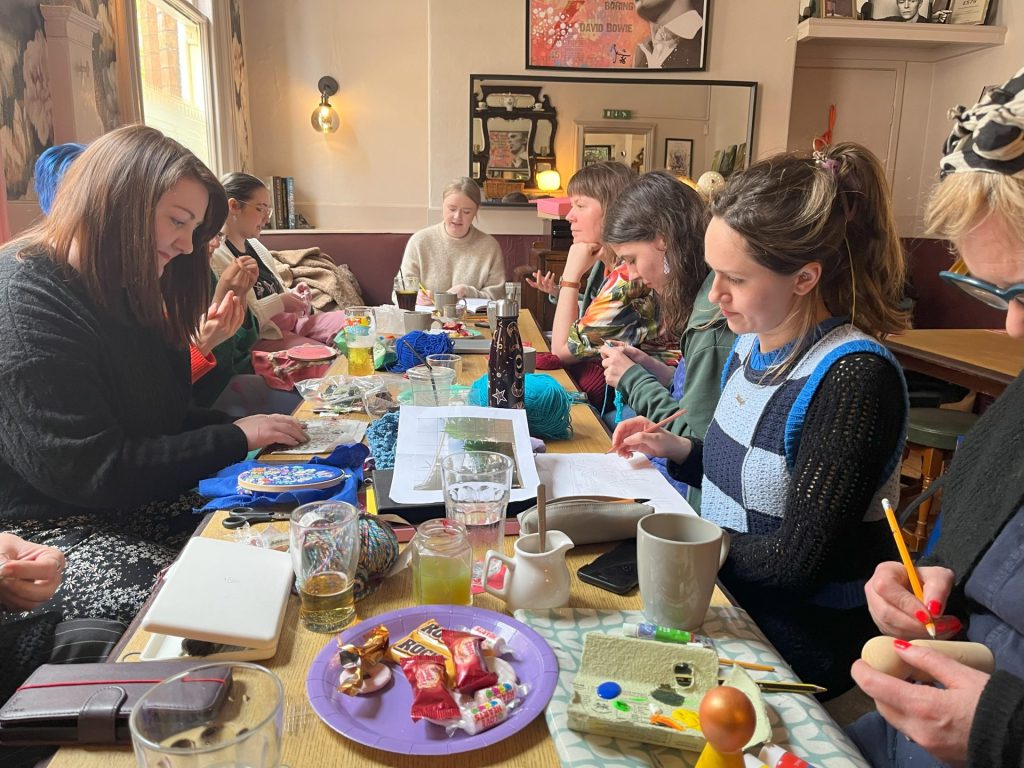 A group of people sit around a cluttered wooden table in a cozy room, engaging in crafts and conversation. Various crafting supplies, snacks, and drinks are scattered across the table. Walls are adorned with artwork and soft lighting.