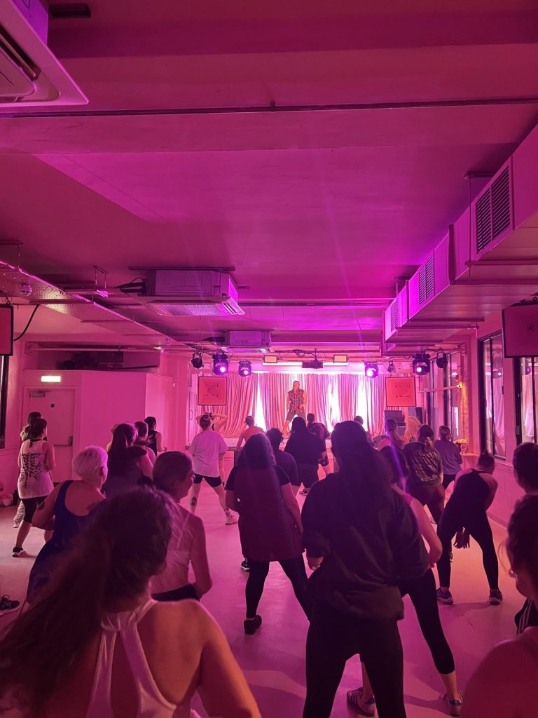 A group of people participates in a dance or fitness class in a room with pink lighting. They face an instructor at the front, and mirrors line the side walls. Ceiling lights highlight the vibrant atmosphere.