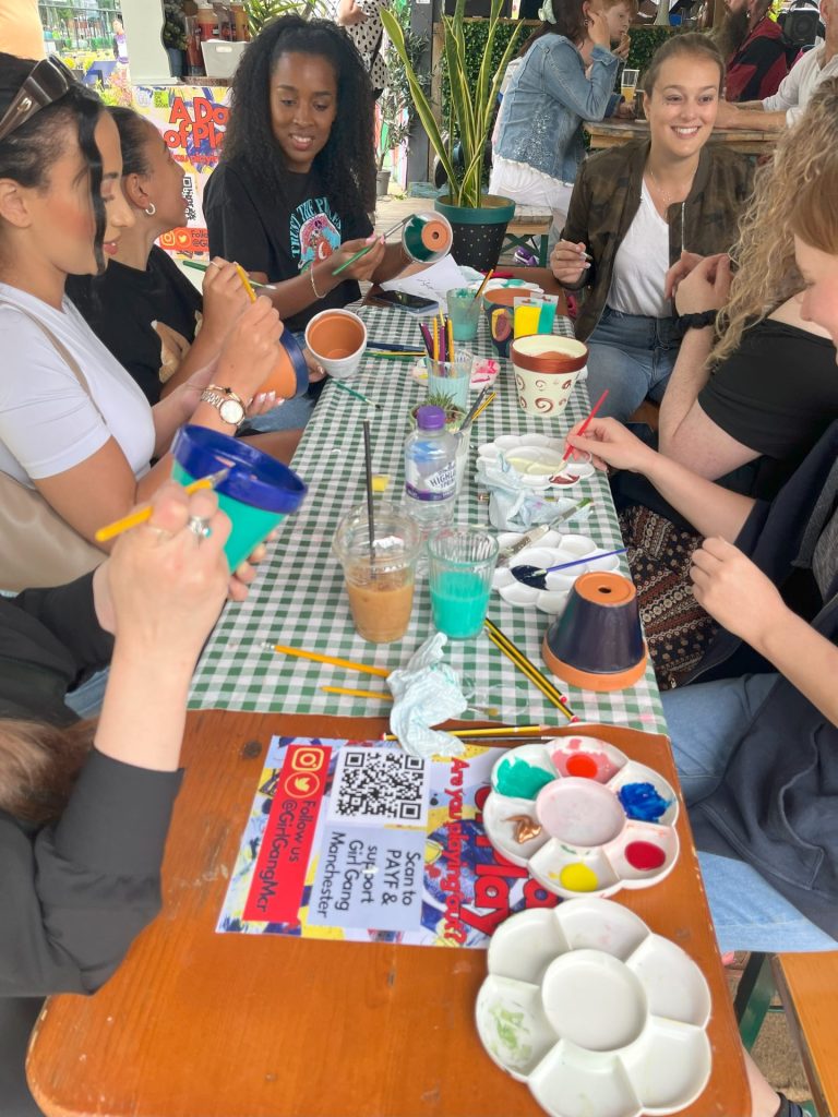 A group of people sit around a table at a pottery painting event. They are painting small pots with brushes, surrounded by paint palettes and art supplies. A checkerboard tablecloth covers the table, and beverages are nearby.