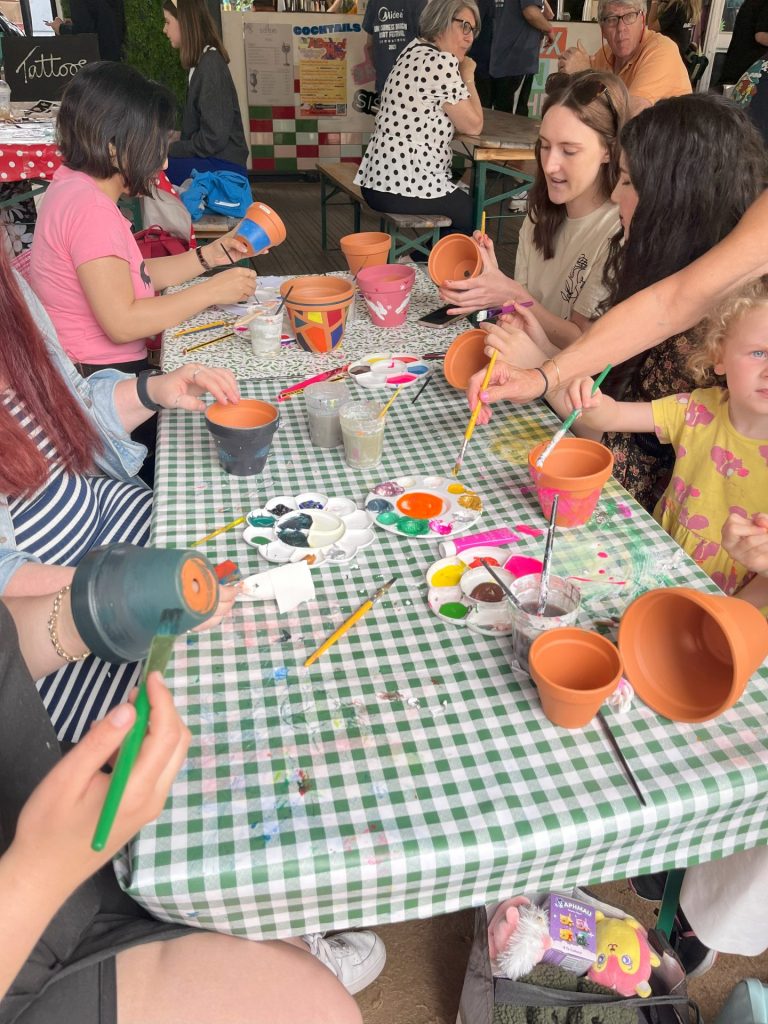 A group of people sitting at a checkered table, painting small terracotta pots with various colors. Brushes and paint materials are scattered on the table. A child is also participating in the activity.