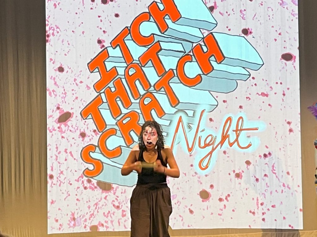 A woman performing in front of a projector screen that reads Itch that scratch, she is looking very outraged