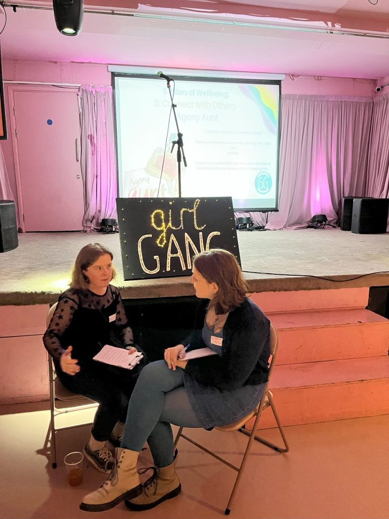 2 people sat on chair talking holding papers and pens, they are sat in front of a stage with a projector screen and sign saying girl gang