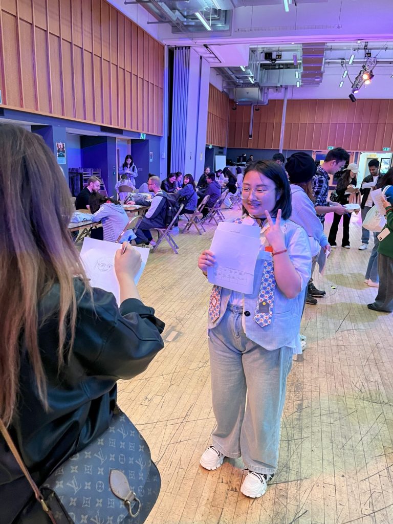 2 people facing each other in a hall full of other people talking in pairs. Person is holding a paper and holding up a peace sign, other person is sketching them