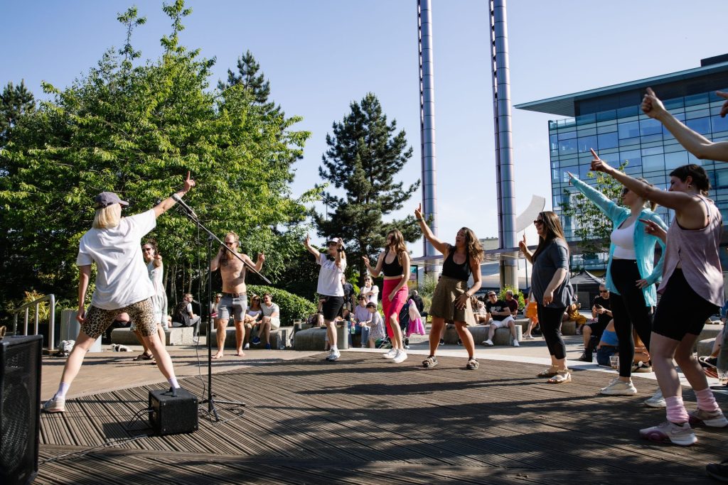 A lively outdoor scene with a group of people dancing energetically on a wooden stage. A performer holds a microphone while others raise their arms. Trees and a modern glass building are visible in the background under a clear blue sky.