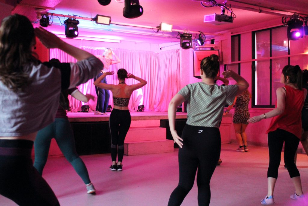 A group of people participating in a dance class on a stage under pink lighting. They are wearing athletic clothing and following the instructor's moves, creating an energetic and lively atmosphere.