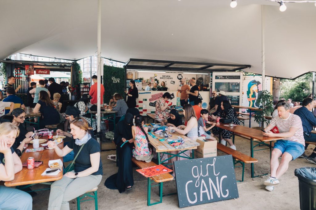 A lively outdoor event with people sitting at wooden tables, eating and chatting. The area is covered by a white canopy. A sign reading "girl GANG" is visible. Stalls and decorations are in the background.