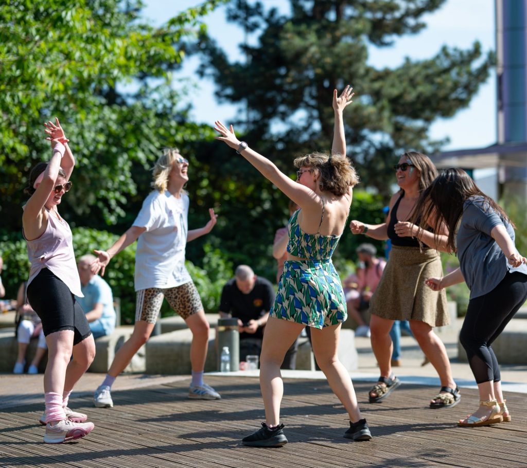 A group of women dancing joyfully outdoors on a sunny day. They are surrounded by greenery and seated people in the background. The women are smiling, with arms raised, and appear to be having a great time.