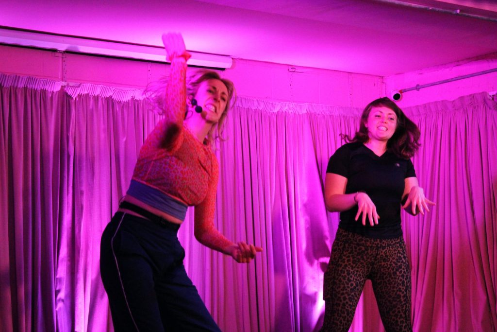 Two people are energetically dancing on a stage with pink lighting. The person on the left is wearing a patterned long-sleeve top and dark pants, while the person on the right is in a black shirt and leopard-print leggings. Curtains hang in the background.