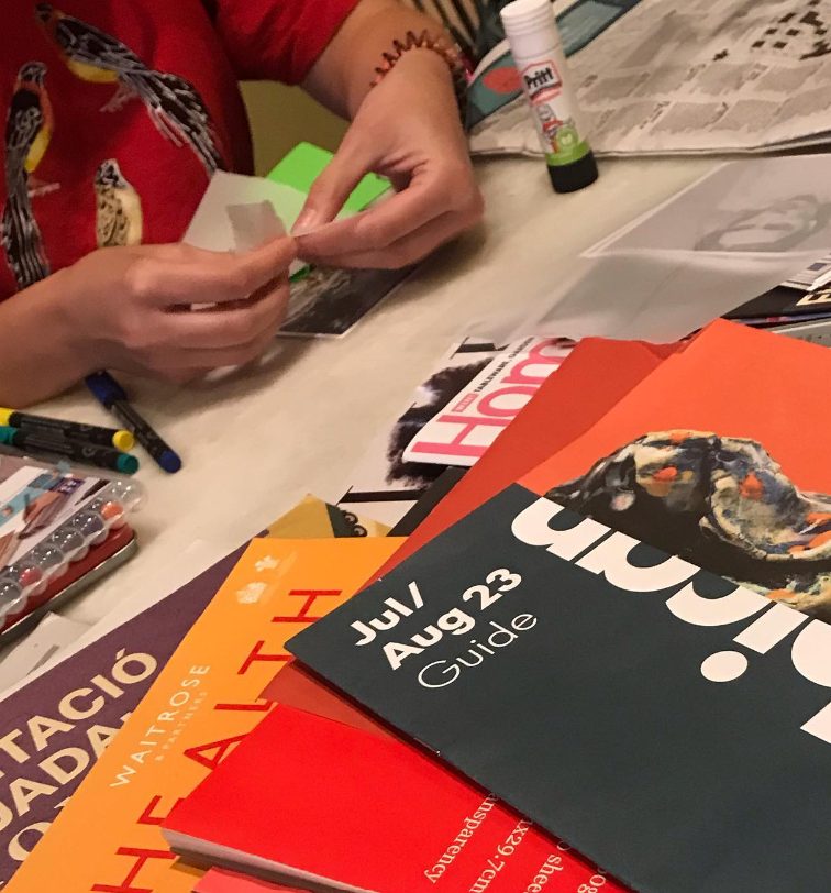 A person in a red shirt is crafting at a table, surrounded by various colorful magazines, scissors, and a glue stick. They are working with paper cutouts.