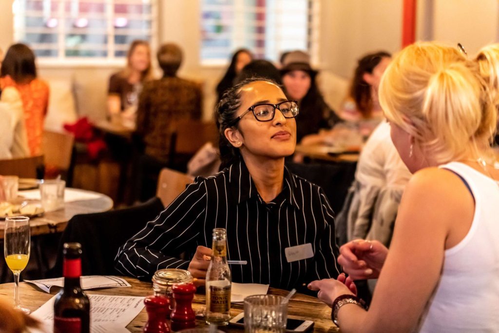 2 people looking at each other in conversation, sat at a table with drinks and papers on the table. other people in the background