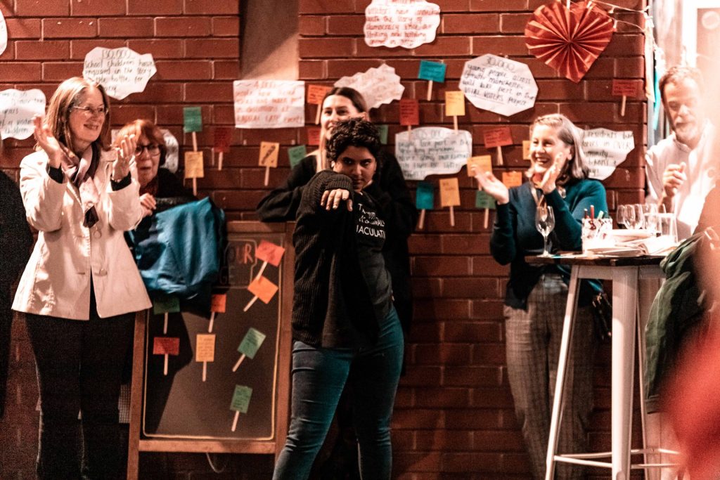 A person stands confidently pointing in front of a board, surrounded by a group of smiling people clapping. The background features a brick wall decorated with colorful notes and cut-out clouds with writing on them.