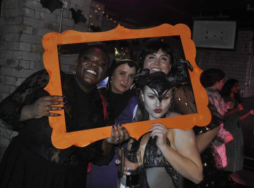 Four people in Halloween costumes pose together, holding an orange picture frame. They stand in a dimly lit room with brick walls, dressed as various characters. One holds a drink, and they all smile or gesture animatedly at the camera.