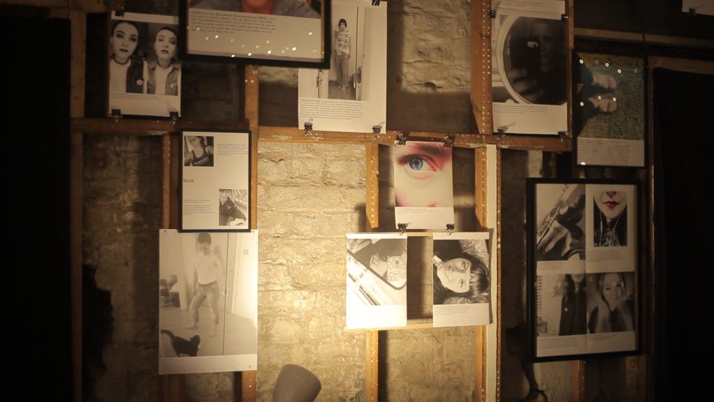 A dimly lit gallery display on a textured wall features various black and white photographs and text panels. One large image highlights a striking close-up of an eye with colorful makeup. The arrangement is artistic and eclectic.