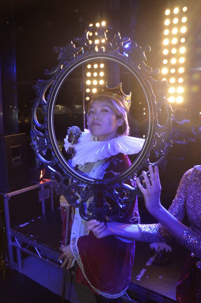 A person dressed in a royal costume, including a crown and ruffled collar, poses with a decorative mirror. Bright stage lights illuminate the background, creating a dramatic effect. Another person holds the mirror frame.