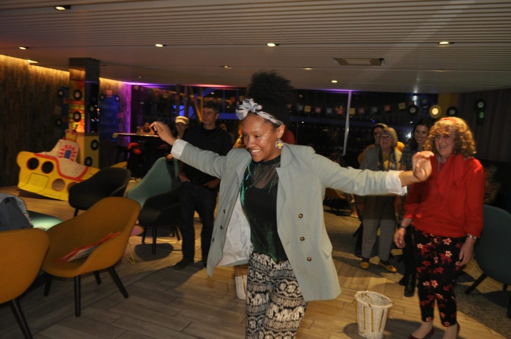 A woman joyfully dances in a lively indoor setting, surrounded by smiling people. The room is decorated with vibrant retro elements and comfortable chairs, creating a fun and relaxed atmosphere.