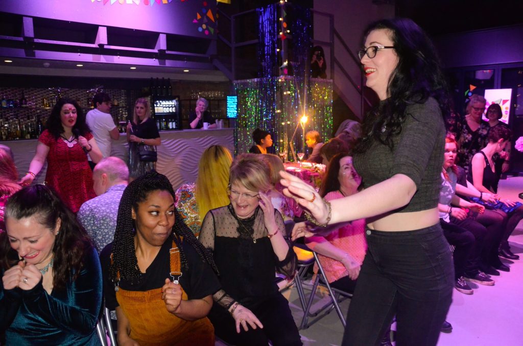 A group of people, both seated and standing, are enjoying a lively event indoors. The room is decorated with colorful lights and streamers. Some people are laughing and clapping, while one person is walking with enthusiasm.